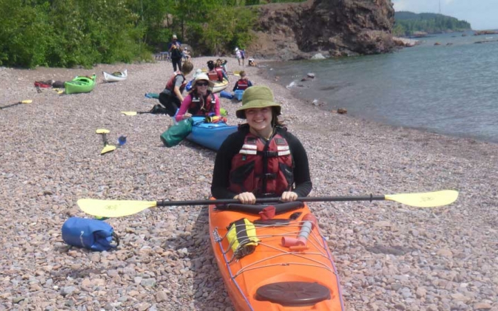 A group of people sit in kayaks beached on the shore of a lake, in an apparent kayaking exercise. 